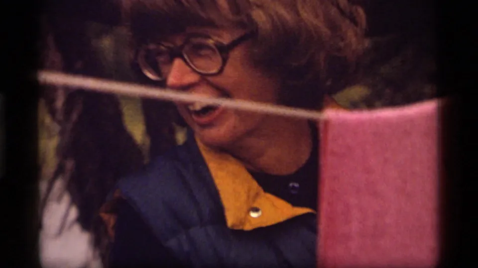 Woman in thick-rimmed glasses and a blue-yellow jacket smiles near a clothesline at the Montana campsite.