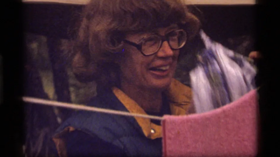 1977 home-movie frame of a woman at a campsite clothesline with a pink towel and trees behind her.