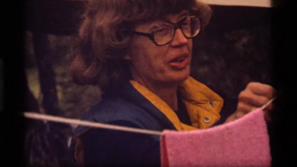 Woman in a blue and yellow jacket appears to speak while handling an item near the campsite clothesline.