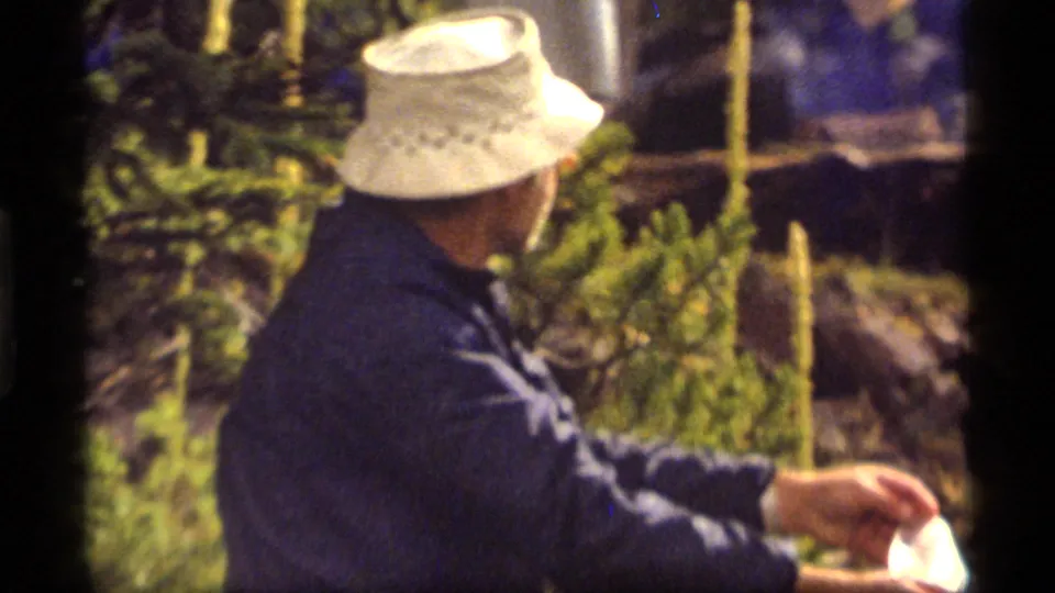 The man in the white hat is shown from the side, reaching toward a white bag in the 1977 Montana home-movie scene.