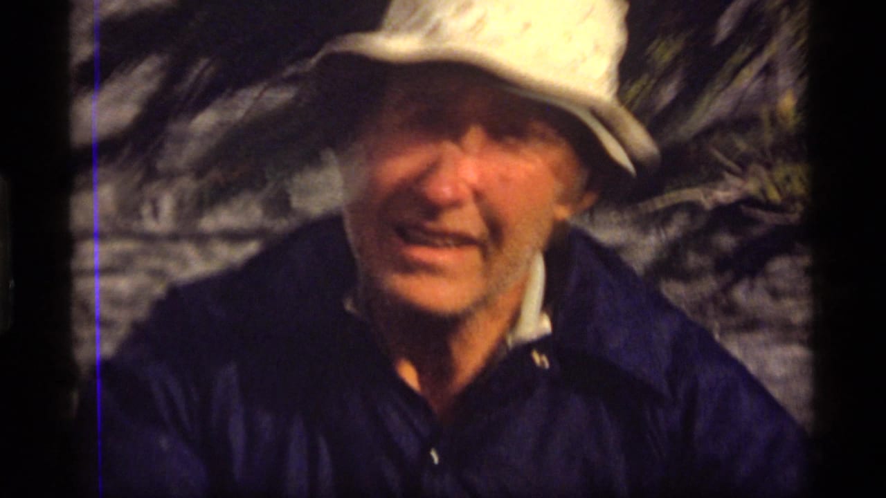 Man in a white hat faces the camera near rippled water and trees.
