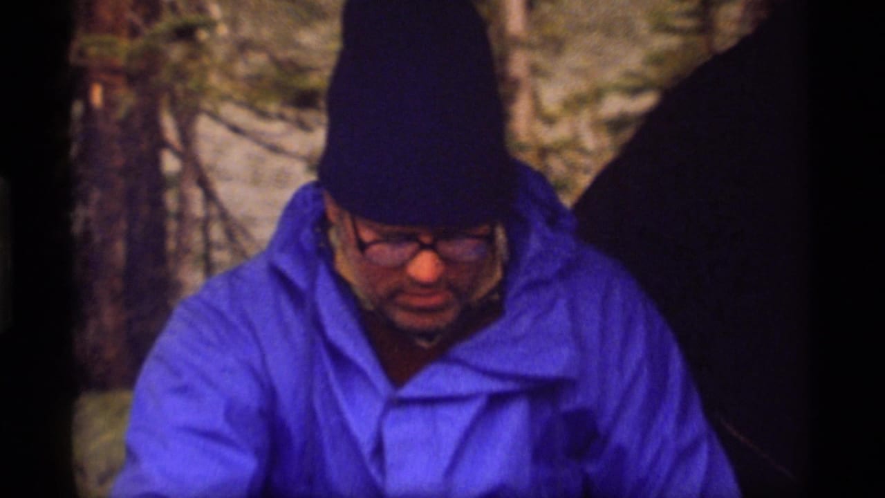 1977 8mm home-movie frame from Sapphire Lake, Montana, showing a man in a blue rain jacket and dark beanie looking down while seated outdoors.