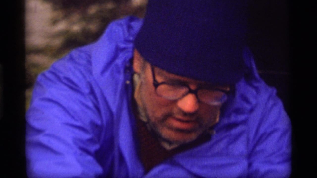 Close view of the man in blue rain gear and glasses leaning forward outdoors.