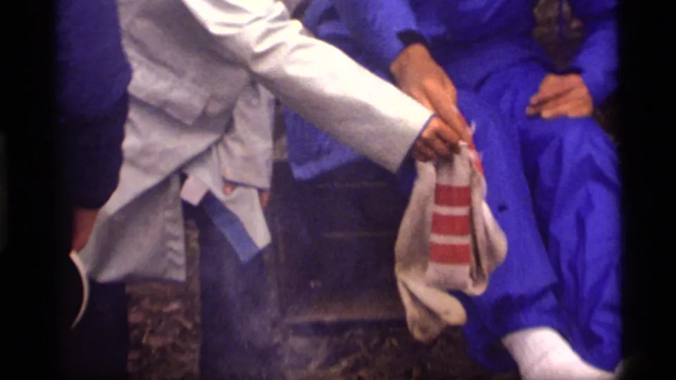1977 Montana camping frame showing a striped sock being handled beside a seated man in blue rain gear, with another person partly visible.