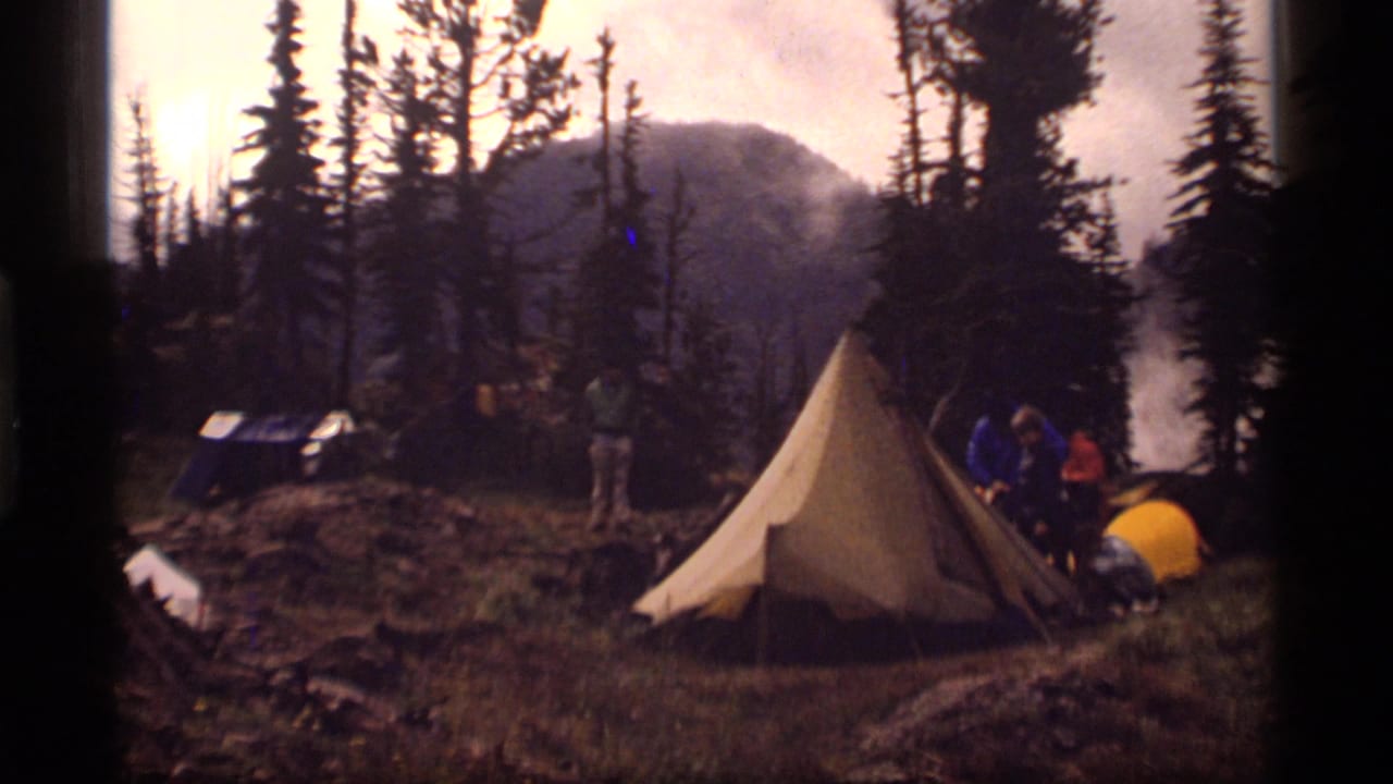 Campsite with an A-frame tent, campers, pine trees, smoke or mist, and mountains in the background.