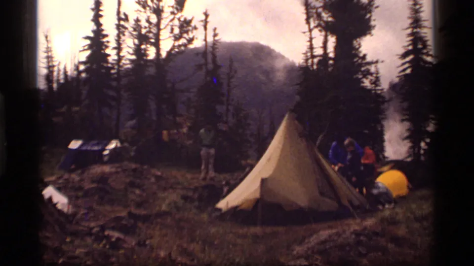 1977 Sapphire Lake, Montana campsite with an A-frame tent, several campers, evergreen trees, smoke or mist, and distant mountains.