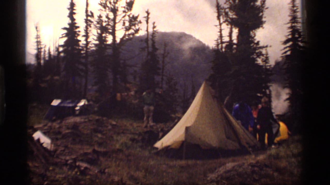 1977 Montana camping scene with an A-frame tent, people near gear, evergreens, and hazy mountains in the background.