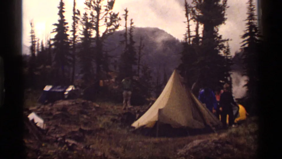 1977 Montana camping scene with an A-frame tent, people near gear, evergreens, and hazy mountains in the background.