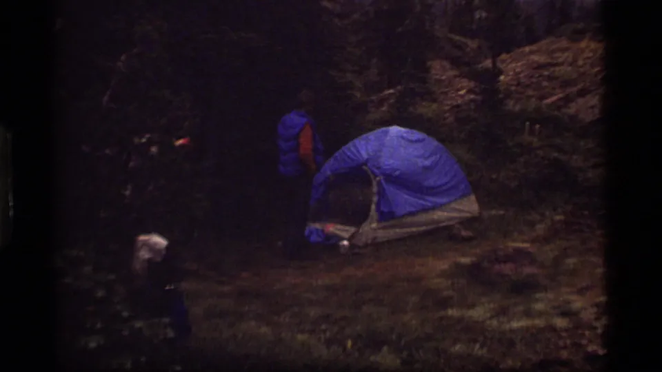 Blue dome tent at a 1977 Sapphire Lake forest campsite, with a camper nearby and rocky, grassy ground around the tent.