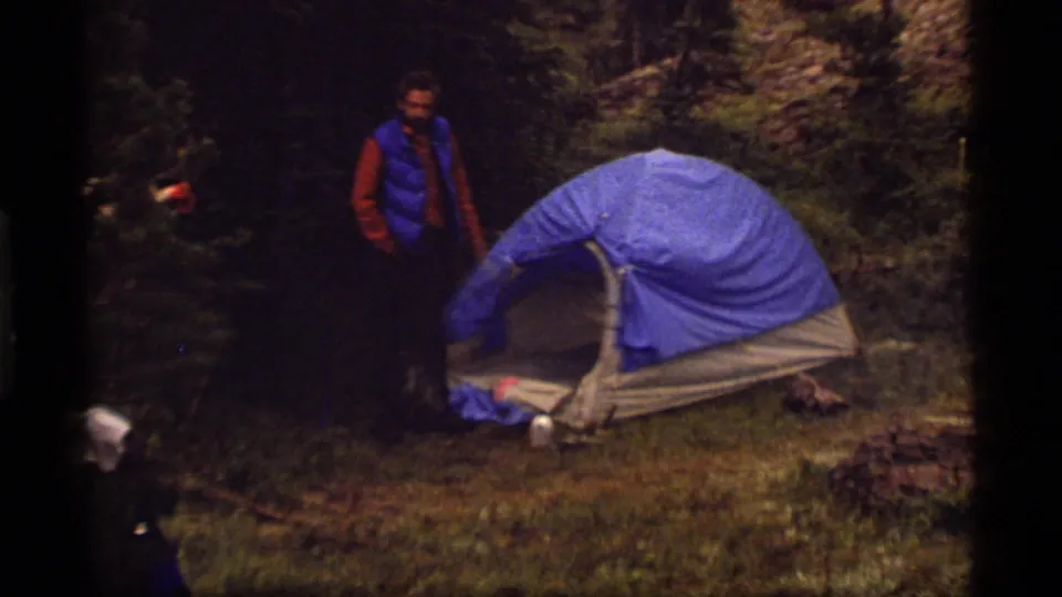 1977 Montana campsite view with a camper in a blue vest beside a blue dome tent, surrounded by trees, rocks, and gear.