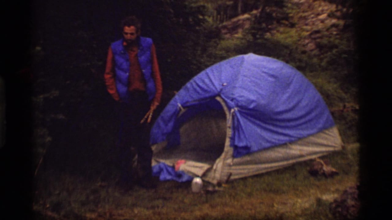 1977 8mm home-movie frame at Sapphire Lake, Montana, showing a man in a blue vest near a blue dome tent in a shaded campsite.