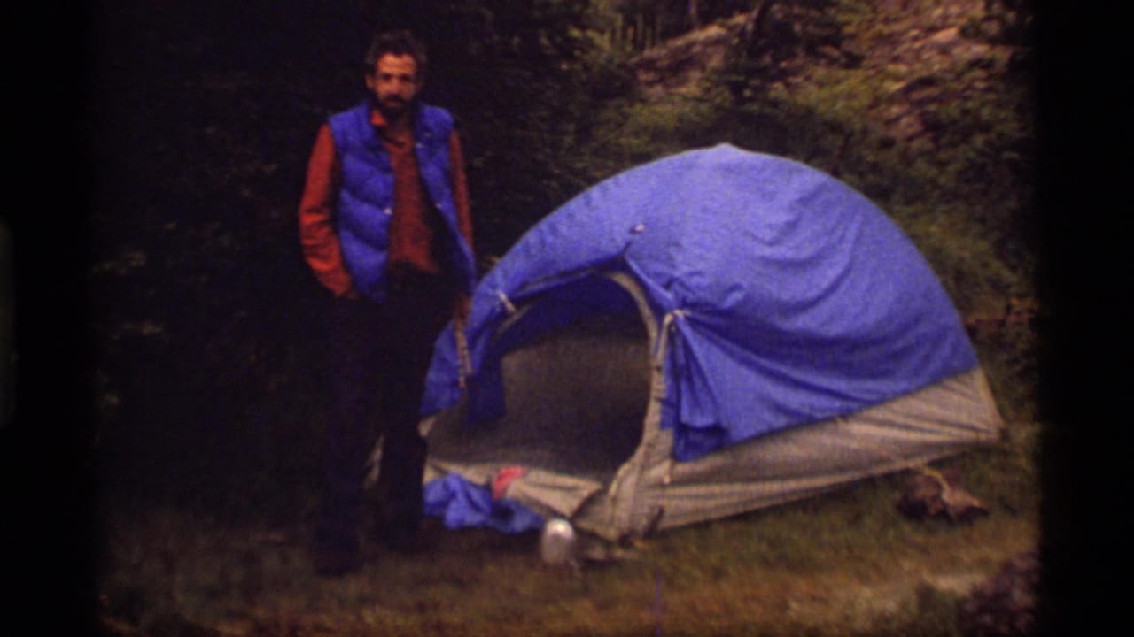 Man stands near a blue tent with wooded ground behind him.