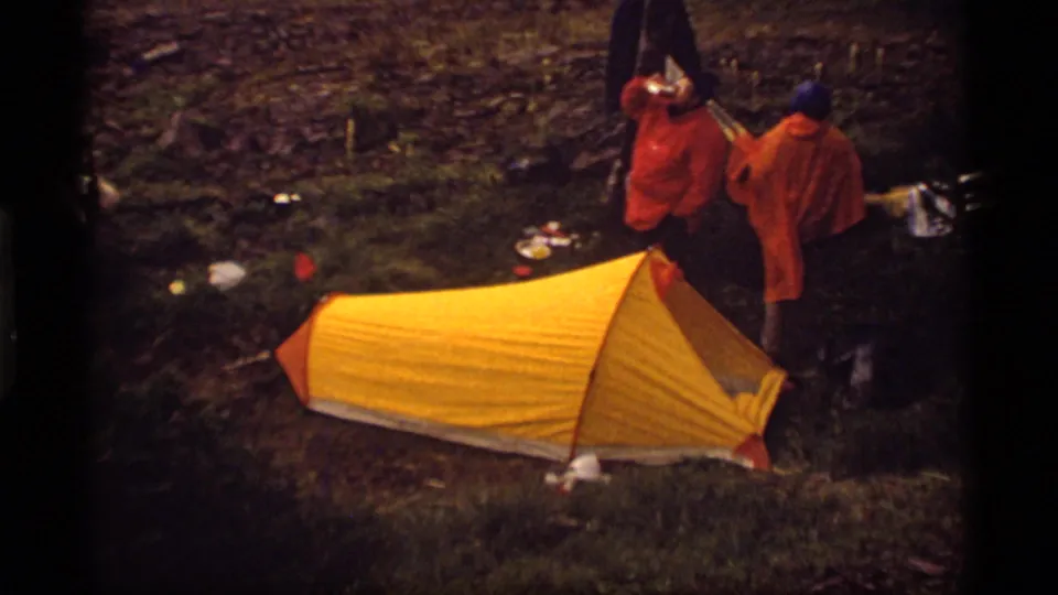 1977 camping footage shows two adults in orange jackets beside a yellow ridge tent, with one person raising a cup near the campsite.