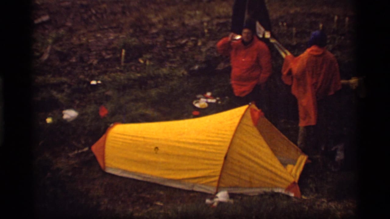 Two men in orange ponchos stand near a bright yellow tent in a forest campsite.