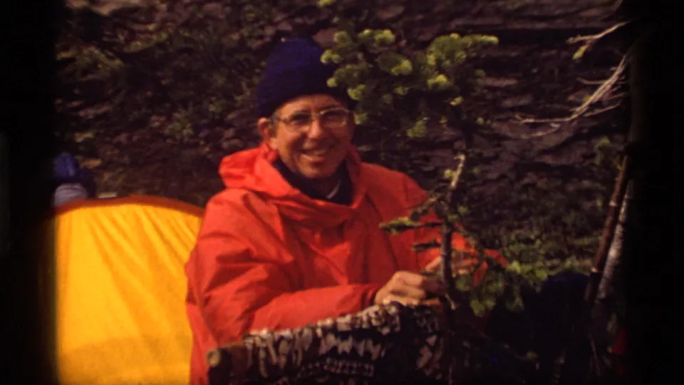 Close 8mm view of a smiling camper in an orange poncho and blue beanie at Sapphire Lake, Montana in 1977.