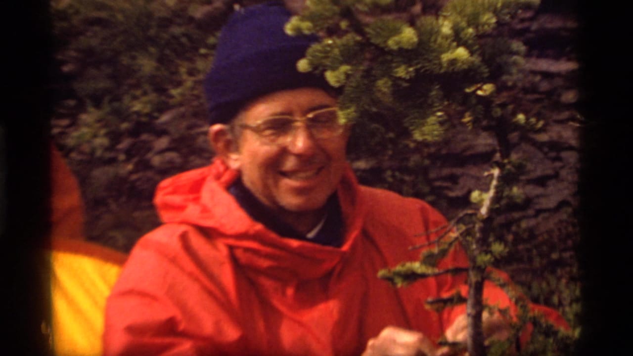 At Sapphire Lake, Montana in 1977, an adult hiker in a red jacket and blue knit cap smiles toward the 8mm camera beside evergreen branches.