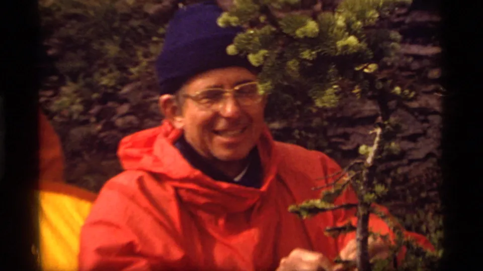 At Sapphire Lake, Montana in 1977, an adult hiker in a red jacket and blue knit cap smiles toward the 8mm camera beside evergreen branches.