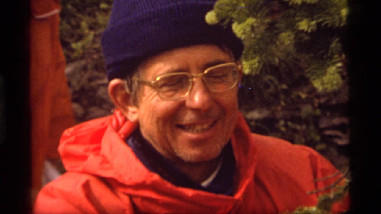 Close 1977 8mm frame of an adult hiker at Sapphire Lake, Montana, smiling in a red jacket, blue knit cap, and eyeglasses.