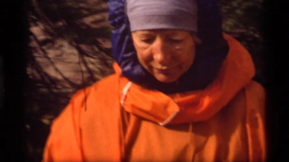 At the campsite, another adult hiker in an orange jacket and striped head covering looks down while standing near dark trees and rock.