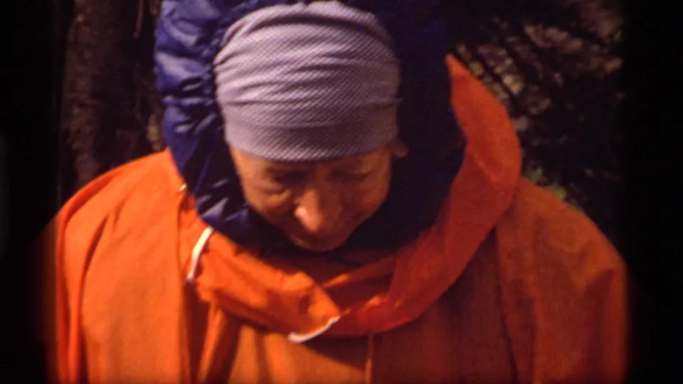 A 1977 Sapphire Lake camping frame shows an adult hiker in orange outerwear looking downward beside trees and rough ground.