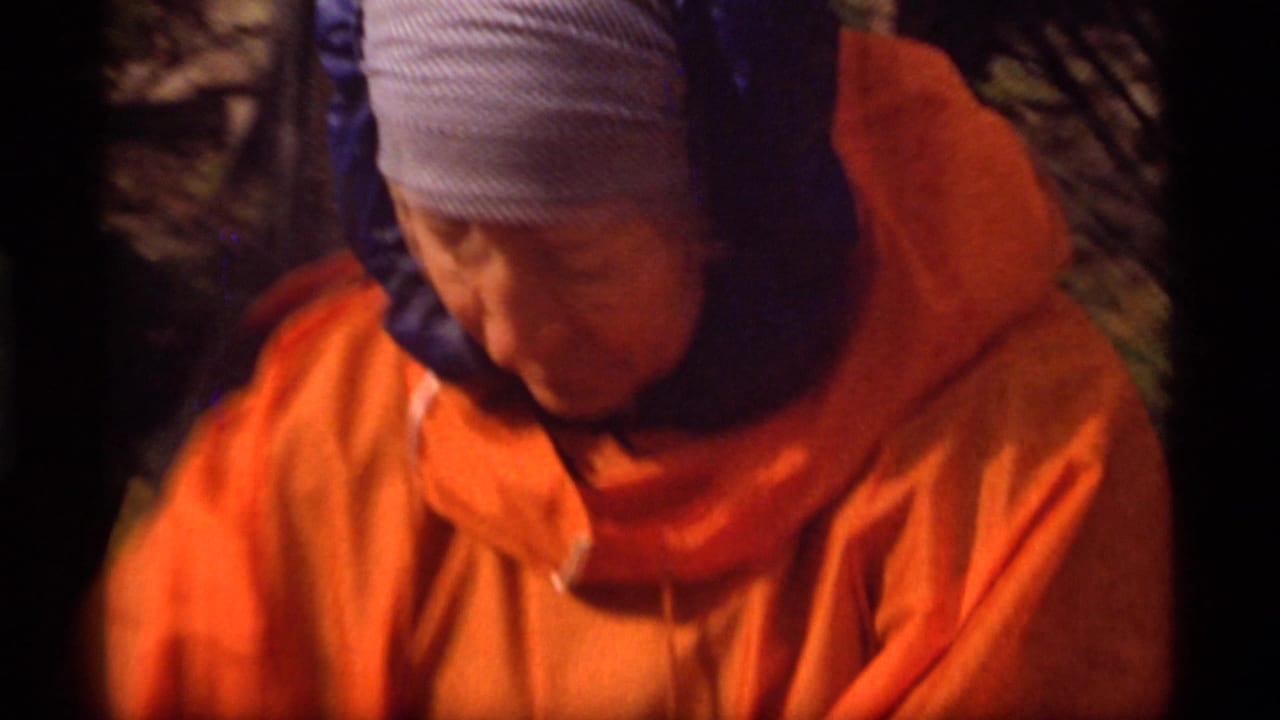 In the supplied 1977 Montana camping footage, a bundled person in an orange parka and blue head covering is seen close up outdoors.
