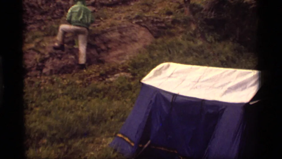 A blue-and-white tent stands on grassy ground while a person in a green jacket climbs a rocky slope in the supplied Montana campsite footage.
