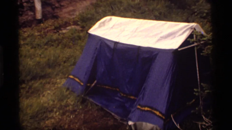 A blue-and-white tent is pitched on grass near brush and trees in the 1977 Sapphire Lake, Montana camping clip.