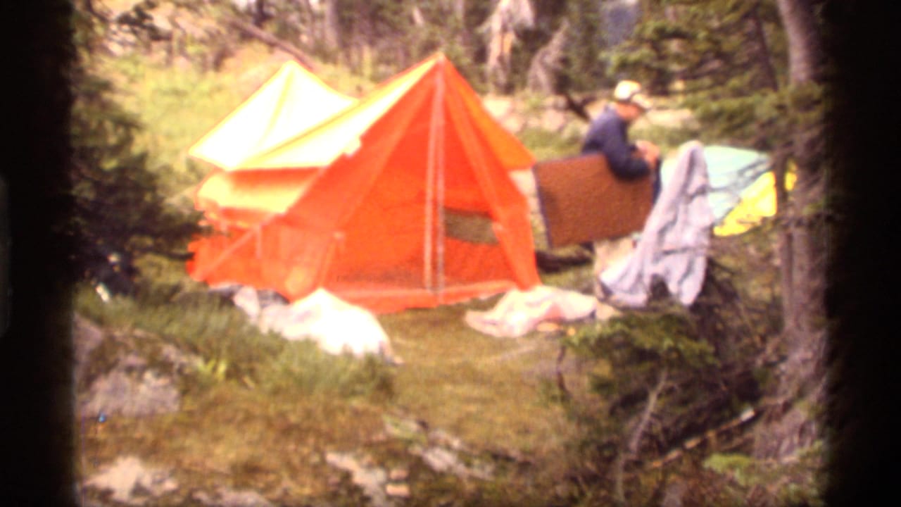 1977 Sapphire Lake, Montana frame showing an orange tent and a man in a hat working with camping gear in a wooded campsite.