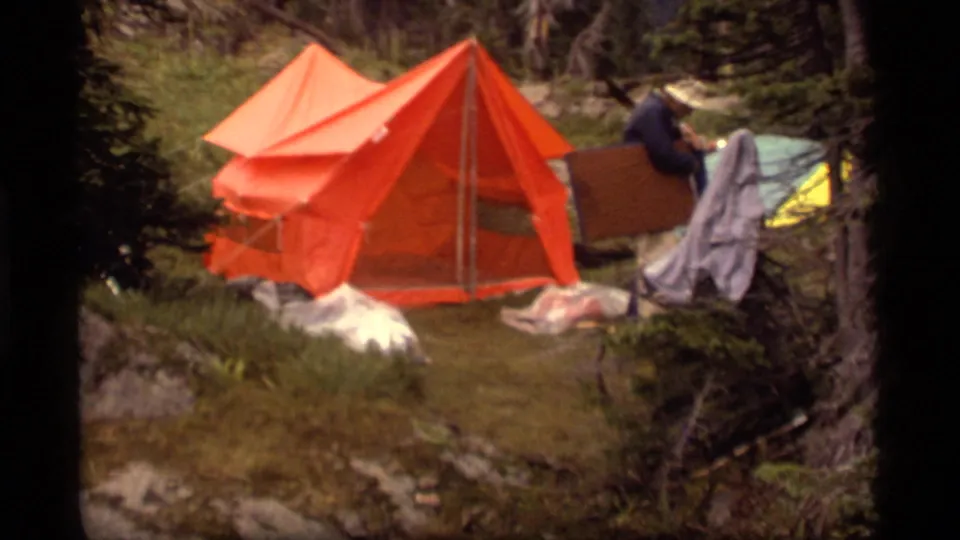 1977 Sapphire Lake, Montana home-movie frame showing an orange tent, scattered blankets, tarps, and a man near gear.