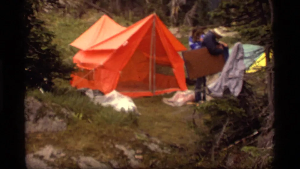 1977 Sapphire Lake, Montana frame showing a man beside an orange tent with tarps, blankets, and a small green tent nearby.