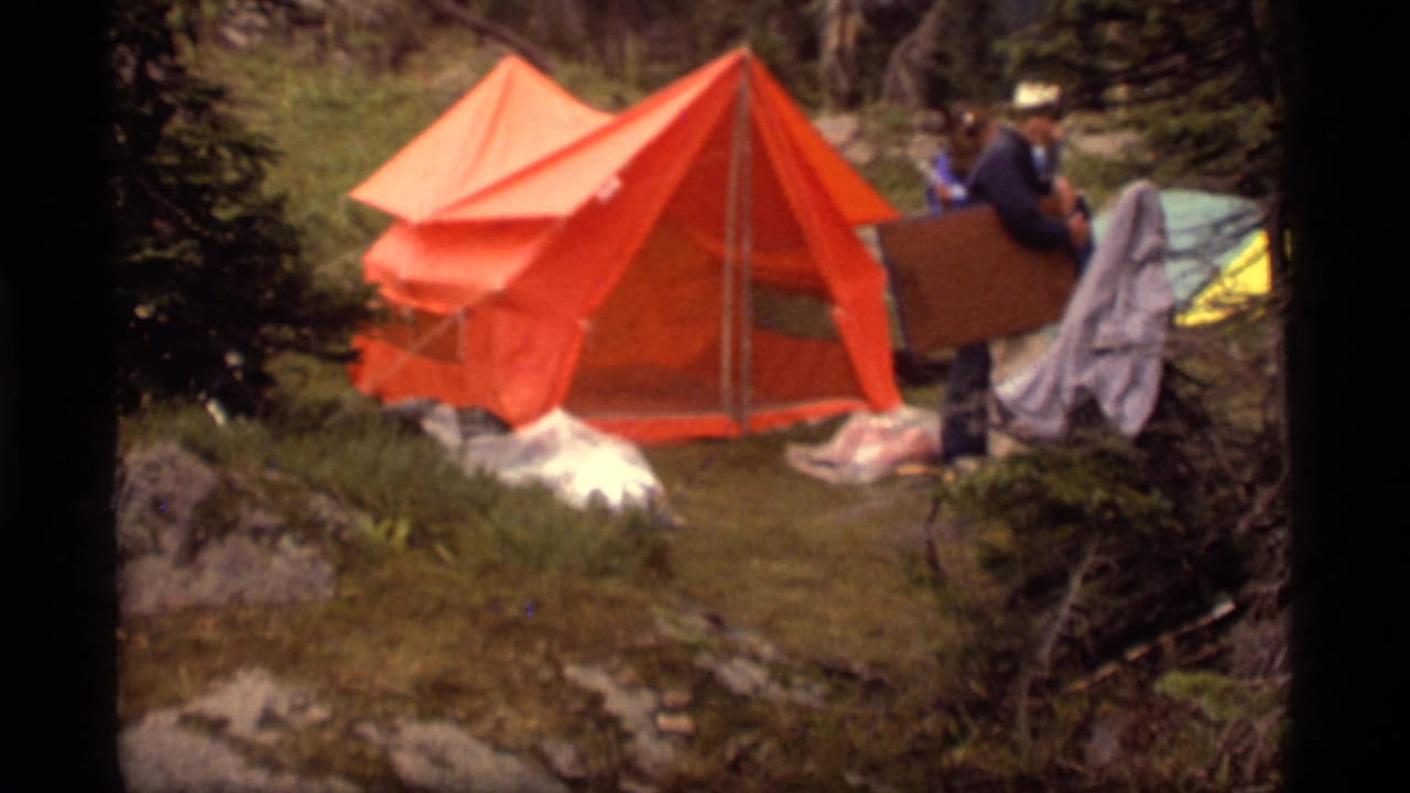 1977 8mm Montana campsite view with a bright orange tent, scattered gear, and a person carrying a box near the trees.