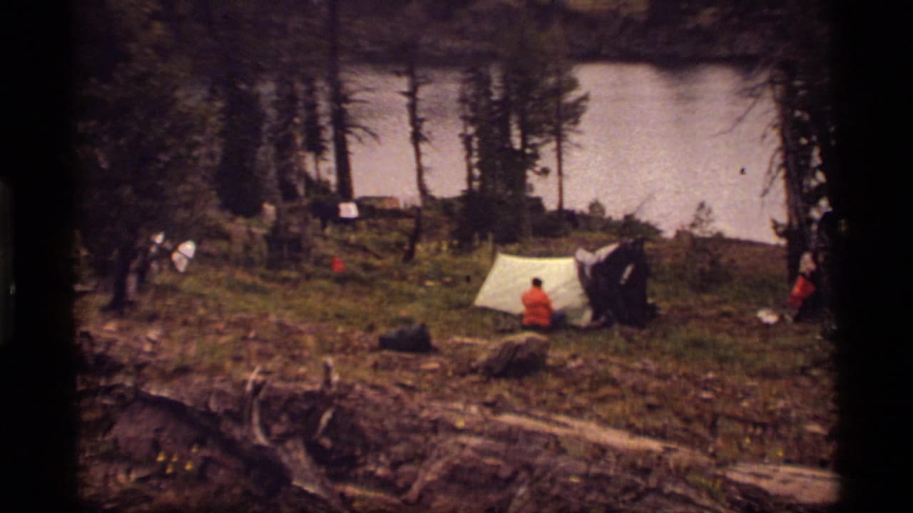 Lakeside campsite with a pale tent, people, trees, and scattered gear.