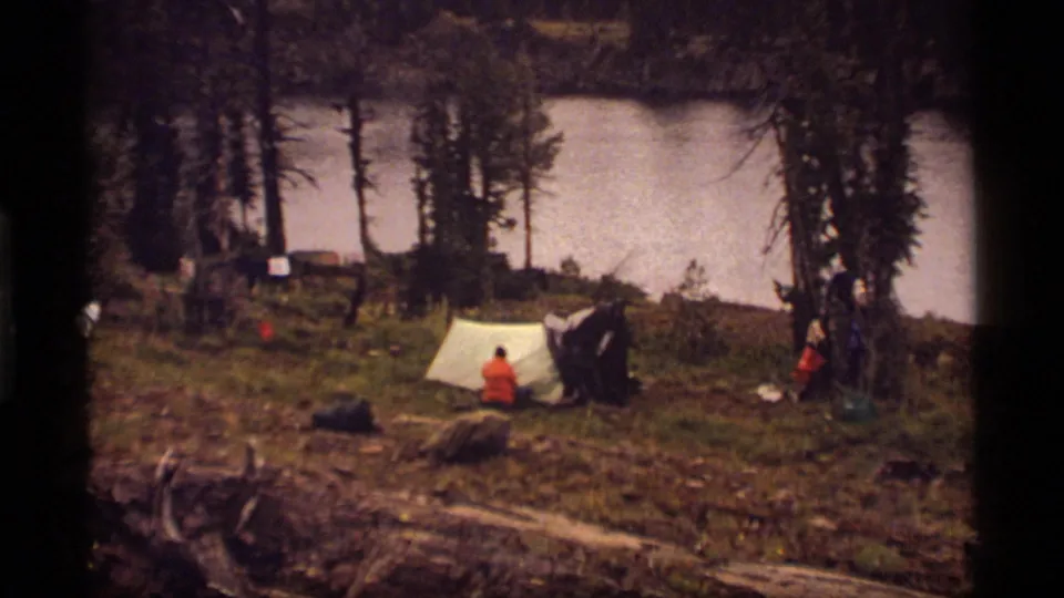 1977 lakeside campsite scene with people near a pale tent and dark shelter, framed by trees and scattered camping items.