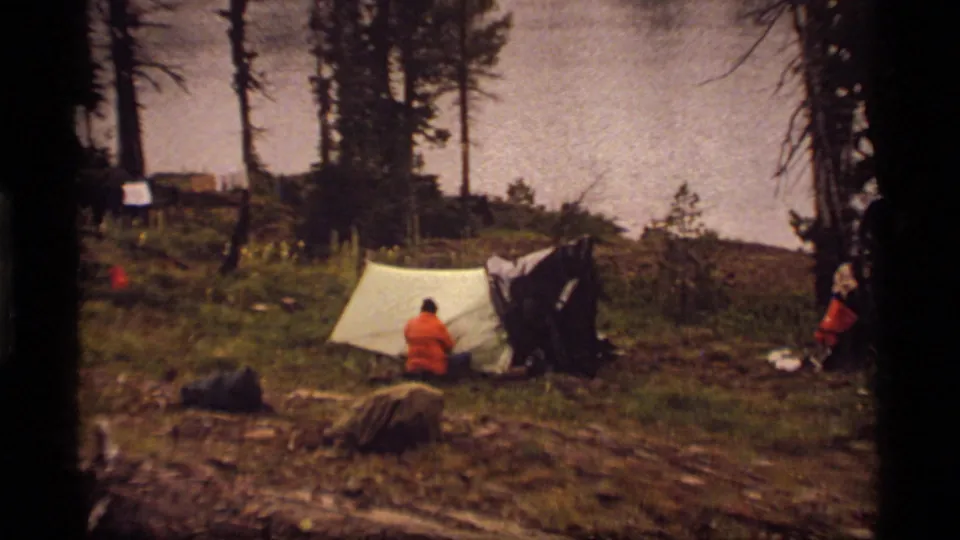 Sapphire Lake campsite view with a person in red near a pale tent, a dark shelter, trees, lake water, and gear in the clearing.