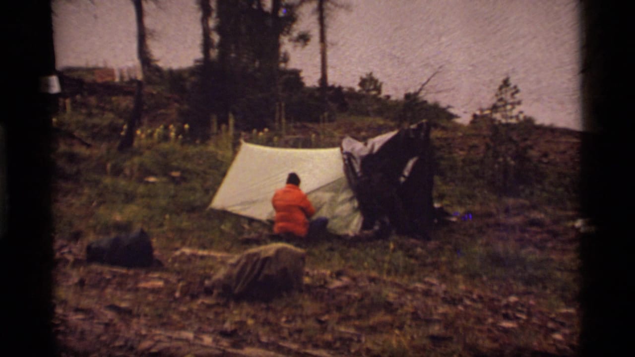 1977 Sapphire Lake camping scene with a person in an orange jacket near a white tent, surrounded by grass, trees, and outdoor gear.