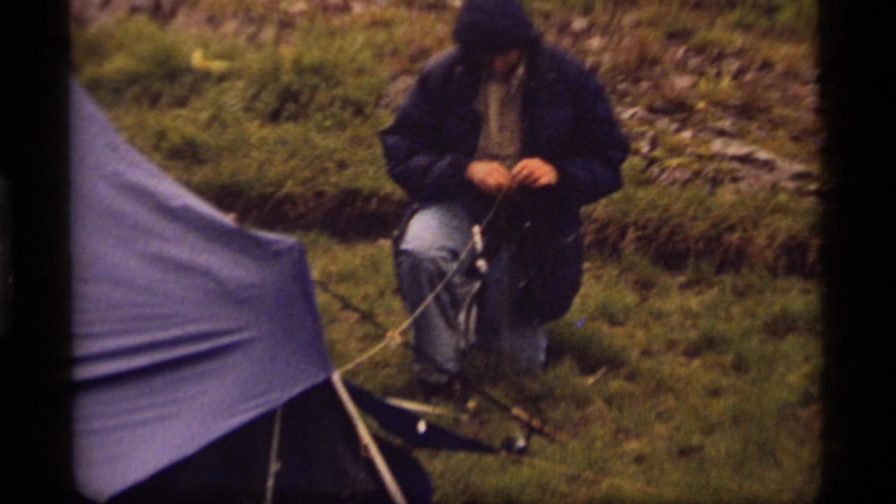 Hooded person kneels beside a blue tarp shelter and adjusts rope or camping gear.