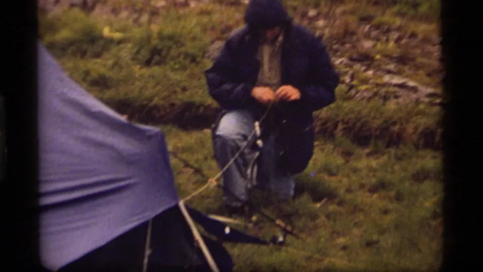 At Sapphire Lake in 1977, a hooded camper kneels beside a blue tarp shelter while working with rope or gear on wet grass.