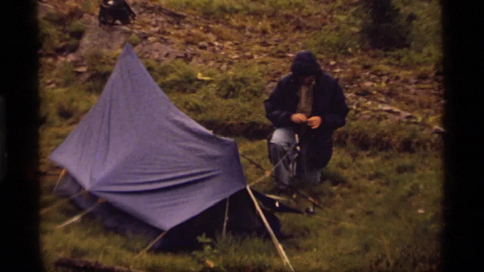A blue tarp shelter dominates the frame as a hooded camper kneels nearby in the 1977 Sapphire Lake camping footage.