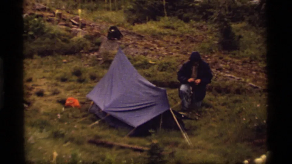 Wide 1977 Sapphire Lake campsite view showing a blue tarp shelter, a hooded camper, an orange item, grass, rocks, and surrounding trees.