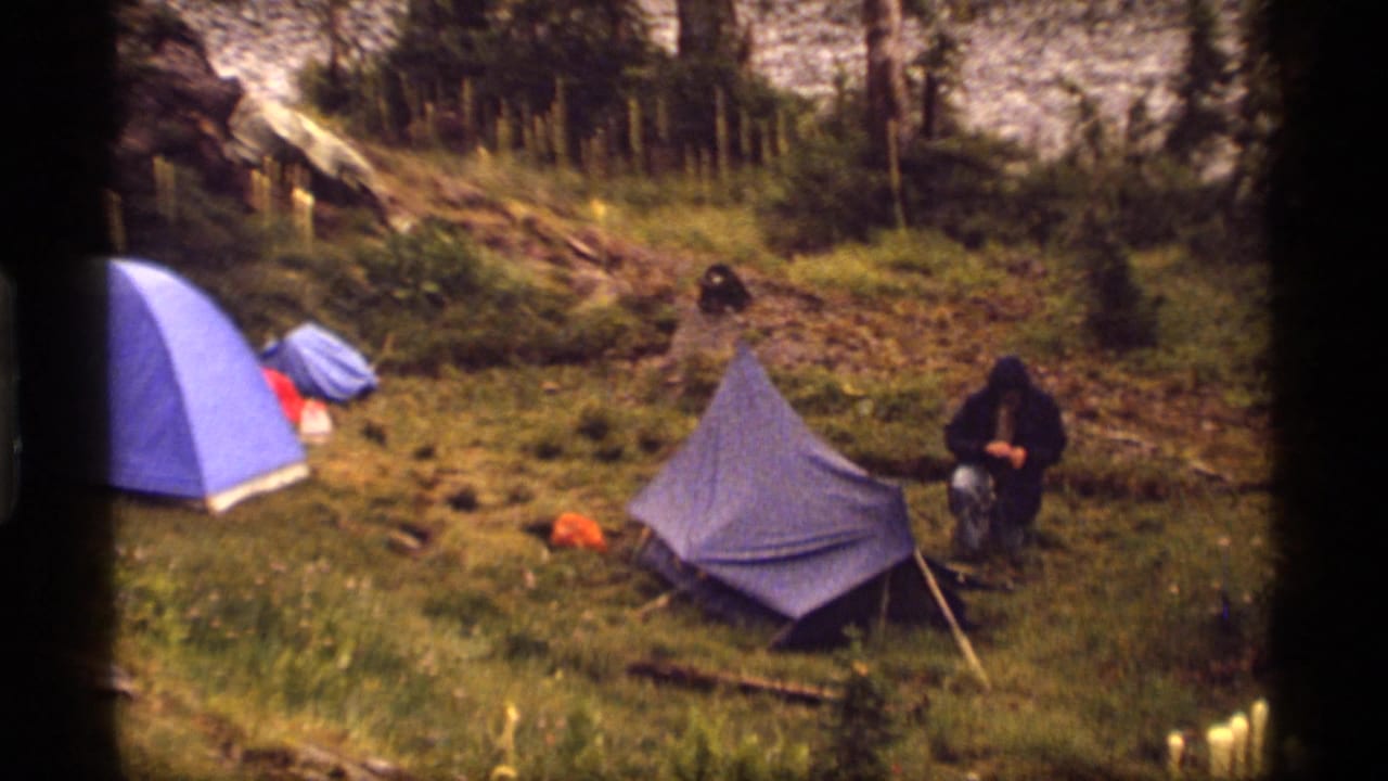 1977 Montana campsite view with two blue tents, scattered gear, and a hooded camper kneeling near the smaller tent.