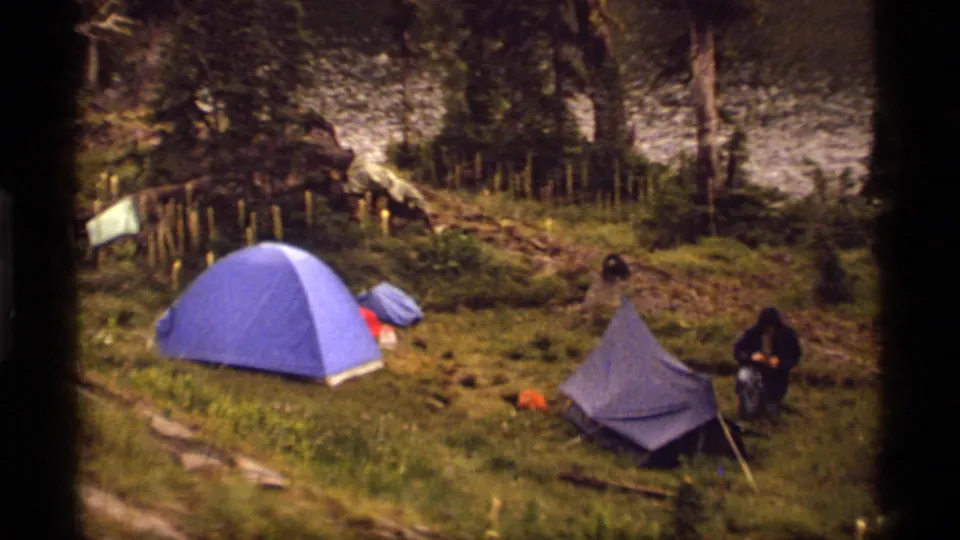 Sapphire Lake camping scene from 1977 showing a blue dome tent, a smaller tent, and a kneeling camper in a wooded clearing.
