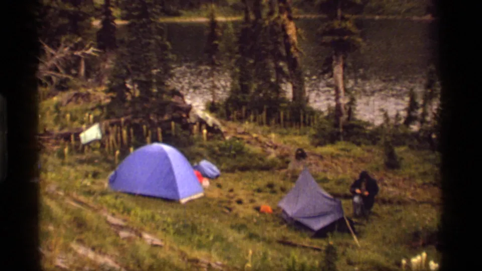 Wide 1977 Montana campsite frame with two blue tents, a hooded camper, gear on grass, pine trees, and water beyond.