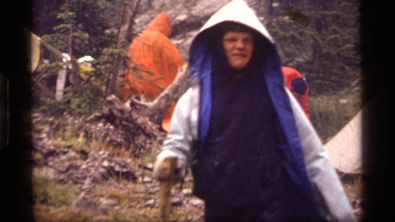 A hiker in blue and white rain covering moves through the wet Sapphire Lake campsite in 1977 footage.