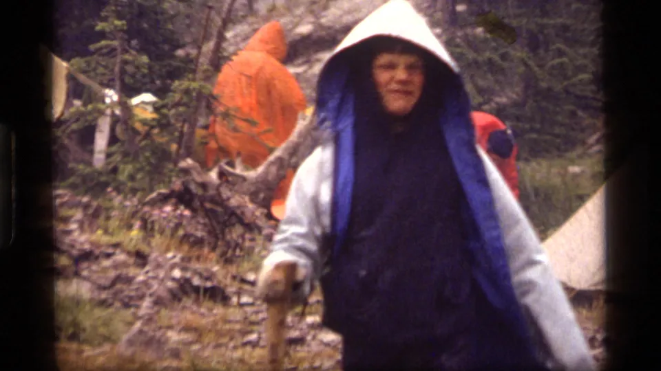 A hiker in blue and white rain covering moves through the wet Sapphire Lake campsite in 1977 footage.