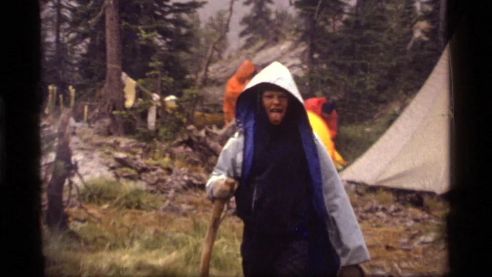 A hiker in layered rain gear holds a stick near a tent at the wet Sapphire Lake campsite.