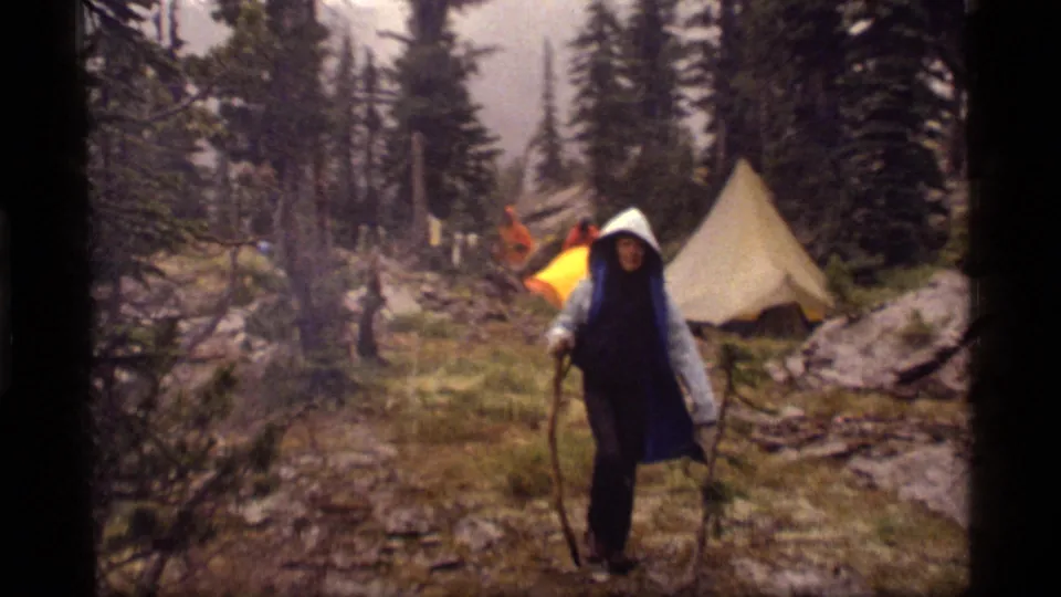 A rain-covered hiker stands on damp ground near a canvas-style tent in the 1977 Sapphire Lake camping footage.