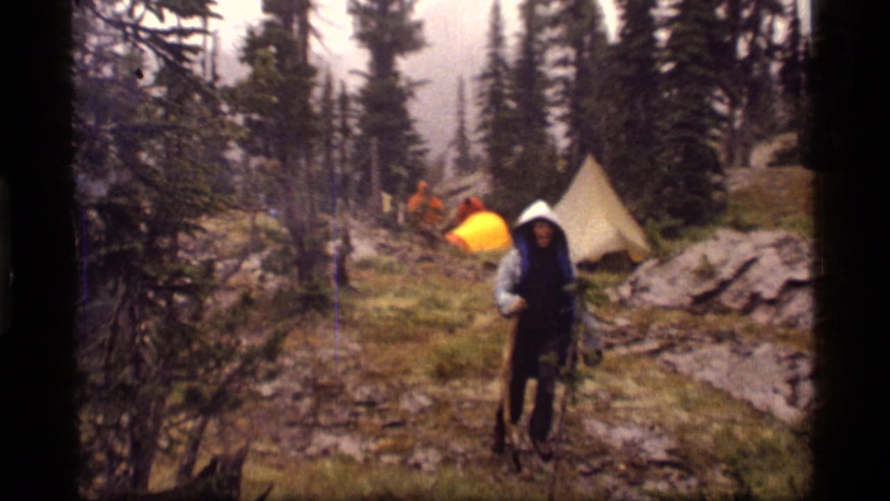 A hooded camper walks through a rainy Sapphire Lake, Montana campsite in 1977, with tents and tall evergreens behind them.