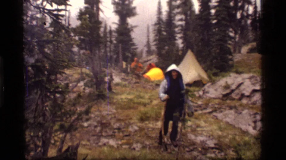 A hooded camper walks through a rainy Sapphire Lake, Montana campsite in 1977, with tents and tall evergreens behind them.