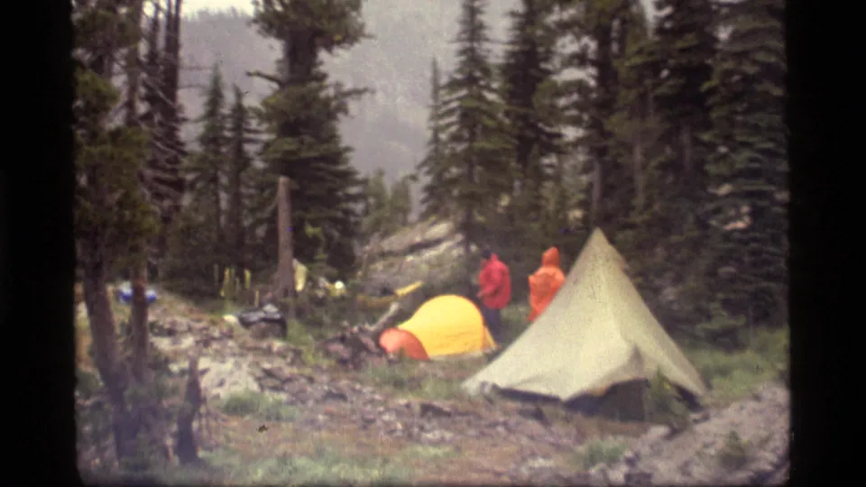 Two campers in bright rain jackets stand near tents at a damp Sapphire Lake, Montana forest campsite in 1977.