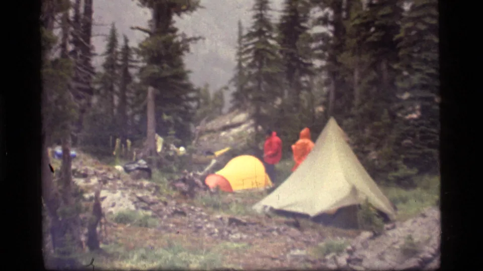Two distant campers in red and orange rain gear stand beside yellow and pale tents in the 1977 Sapphire Lake forest campsite.